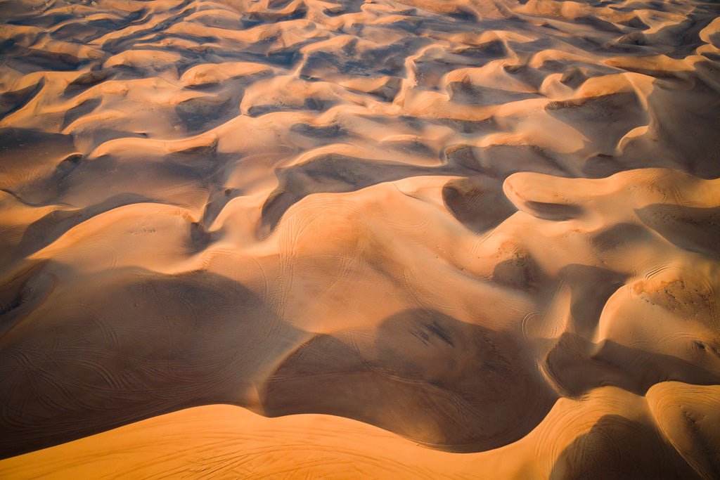 The "Dune Part IV Colour" photograph by FN Prints showcases a breathtaking aerial view of the UAE desert. It features an expansive landscape covered in undulating golden-orange sands, where the sun casts shadows that create an artistic pattern of light and dark areas. The dunes appear smooth, with some tire tracks visible, adding to the serene and vast scene.