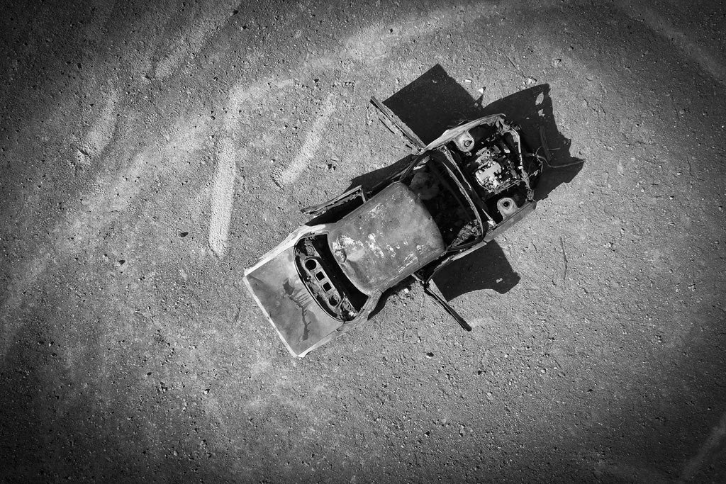 Echoes of Abandonment by FN Prints captures a black and white aerial photograph of an abandoned, deteriorated car shell lying on a deserted, rough surface. The burnt-out vehicle is stripped of its wheels, windows, and most components, leaving only a rusty skeletal frame. Dust and light tire marks surround this compelling monochromatic artwork.