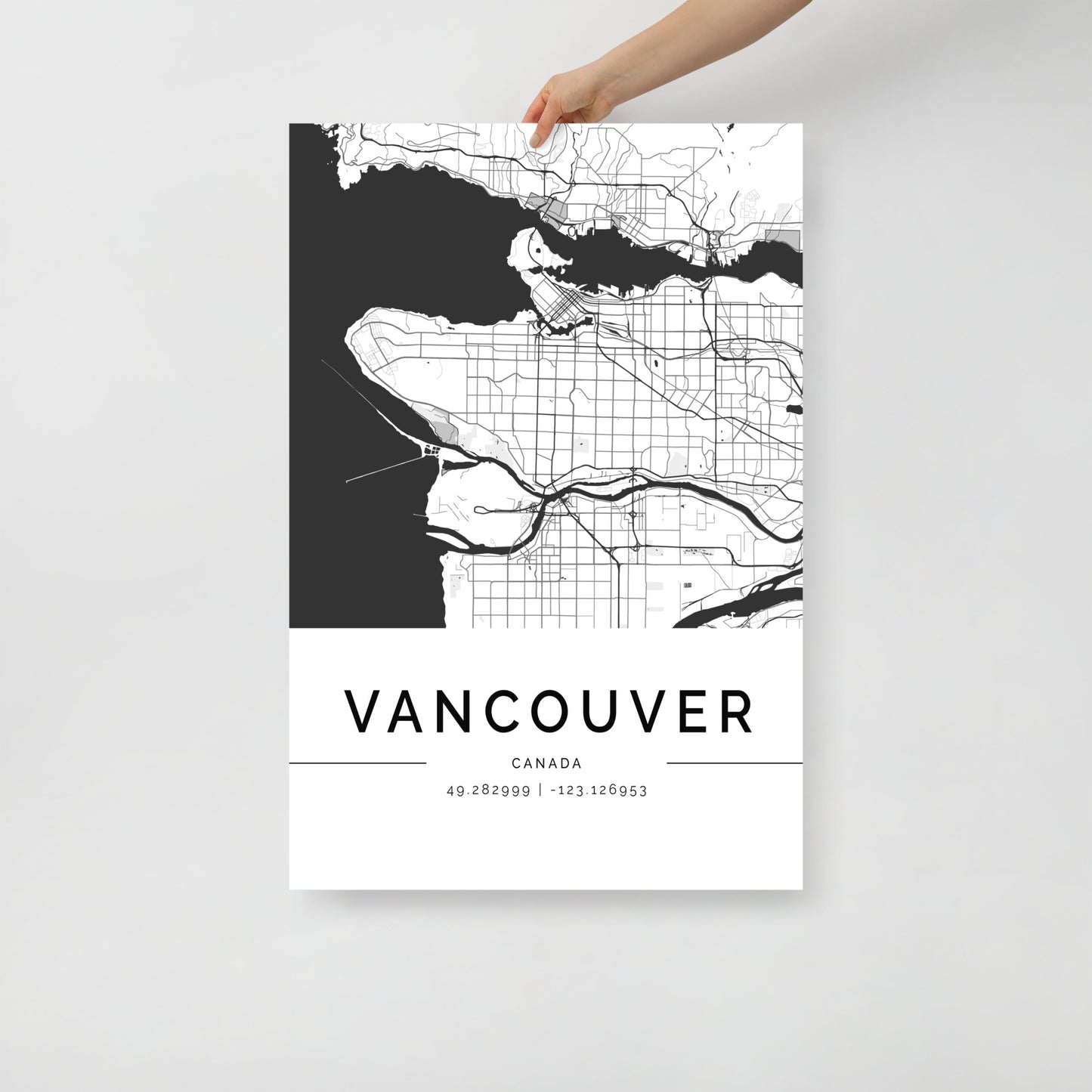 A person holds an FN Prints' Vancouver Map poster, its minimalist black and white design elegantly capturing the cityscape with streets and water bodies. The precise geographic coordinates, 49.282999, -123.126953, are displayed prominently on this striking piece of wall art against a plain backdrop.