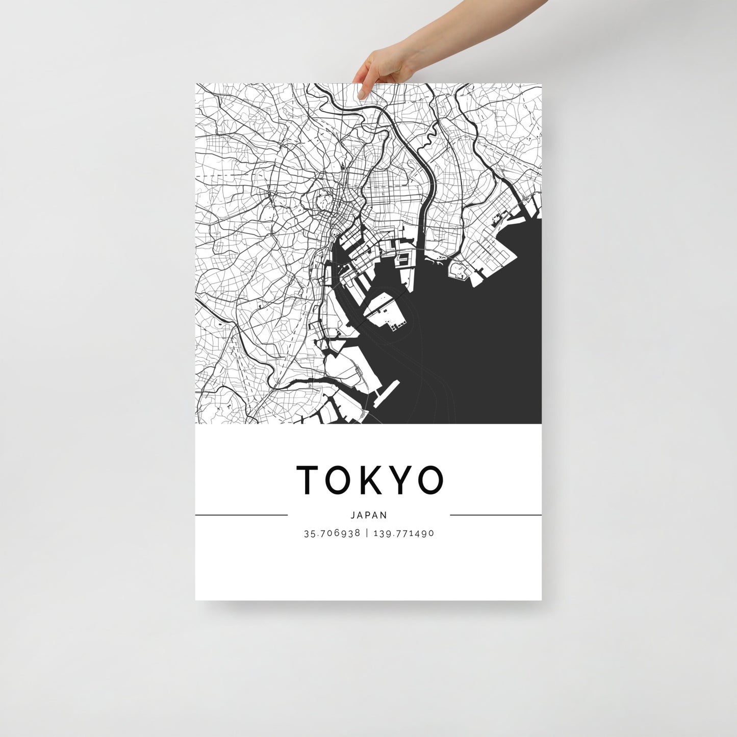 A person holds an FN Prints Tokyo Map featuring a minimalist black and white design of city streets and waterways. This striking wall art includes the geographic coordinates 35.706938 | 139.771490, encapsulating the essence of iconic landmarks in Japan's vibrant capital.