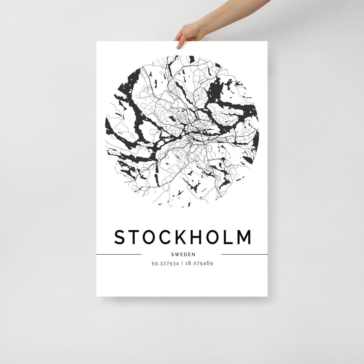 A hand displays a high-quality paper map print from FN Prints, featuring a circular black and white map of Stockholm, Sweden. At the bottom, this Stockholm Map wall art tastefully includes the city's name and its geographical coordinates.