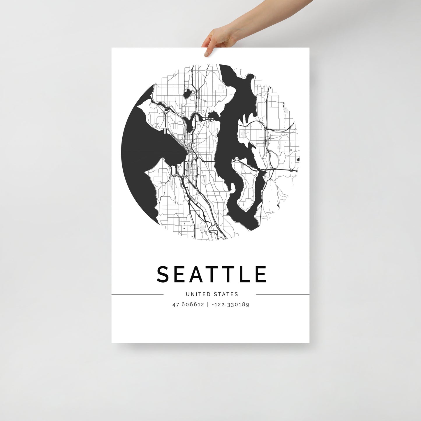 A hand holds a minimalistic black and white Seattle Map poster by FN Prints, crafted on premium-grade paper. This urban landscape artwork showcases a circular depiction of the city's streets and water bodies, with the precise coordinates listed below: 47.606612, -122.330189.