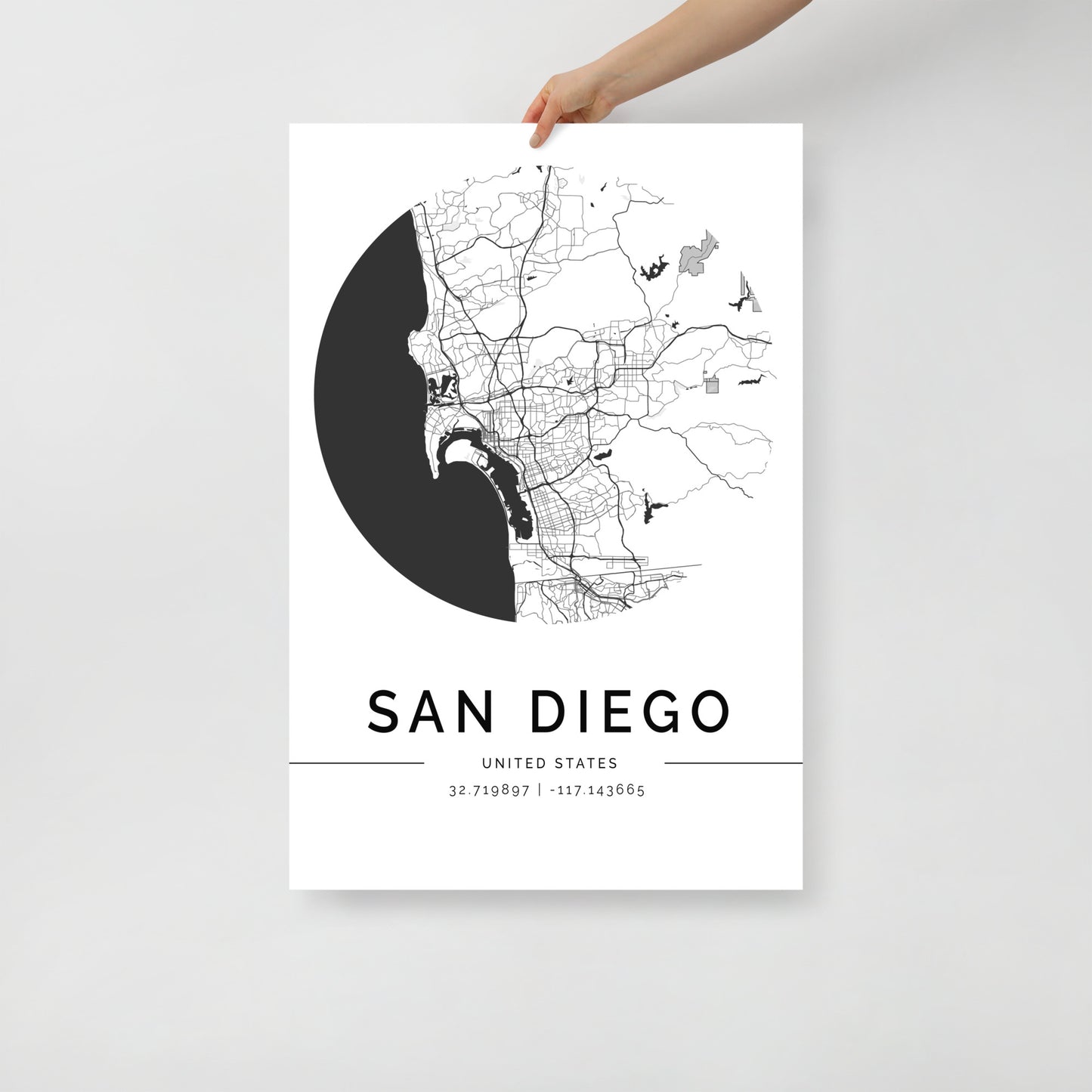 A person is holding the FN Prints' San Diego Map, a minimalistic black and white poster showcasing the city's streets and geographical coordinates (32.719897, -117.434965). The circular design stands out against a white background, making it an ideal piece of coastal wall art for adding an elegant touch to your decor.