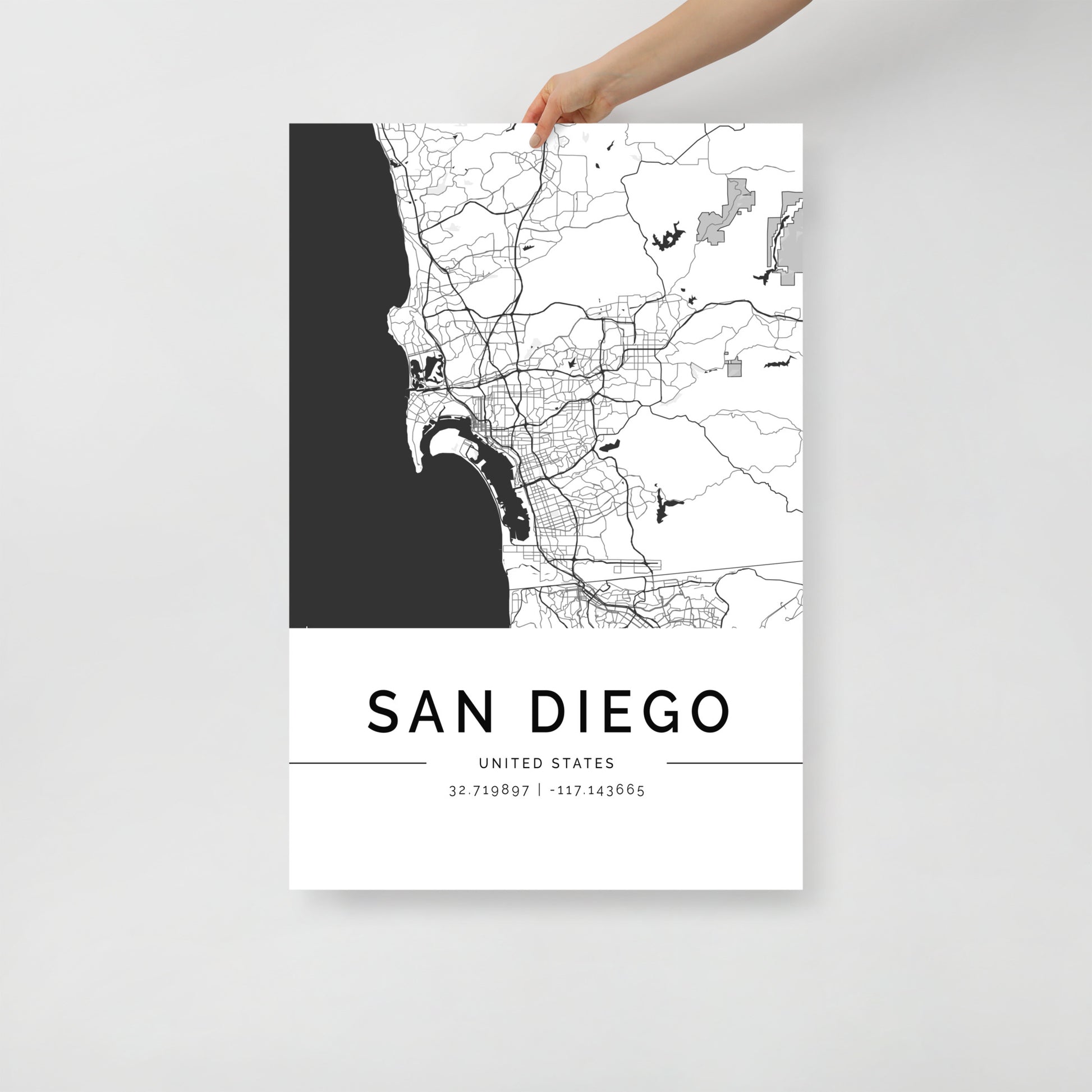 A person holds the FN Prints San Diego Map, featuring a striking black and white depiction of the city's layout. This minimalist design serves as coastal wall art, elegantly displaying geographic coordinates at the bottom for an added touch of sophistication to any space.