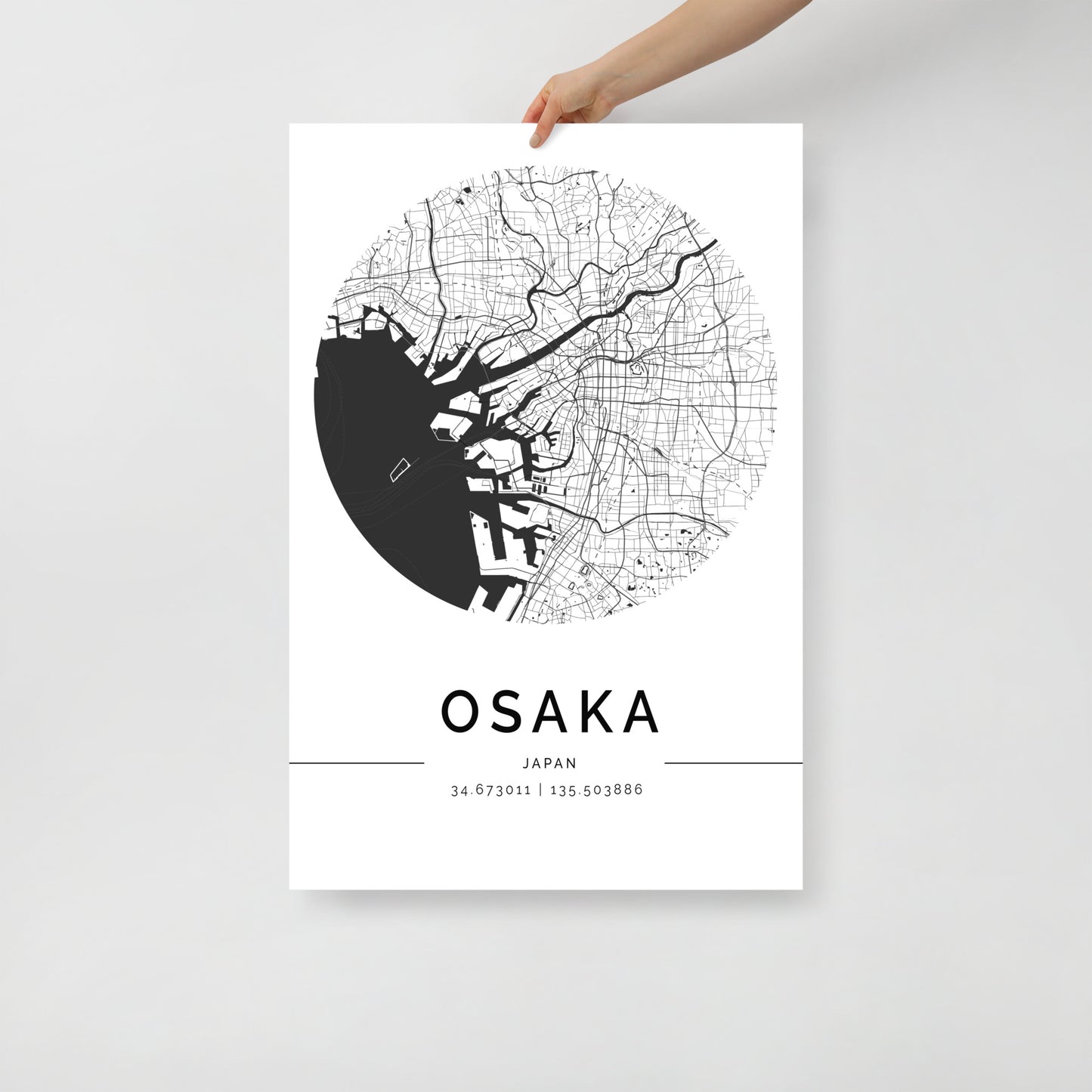 A person's hand holds a stunning piece of art created by FN Prints, the Osaka Map, a minimalist black and white poster that features intricate street lines and the lively Osaka harbor. This elegantly modern design includes coordinates (34.673011, 135.503888) for an added touch of precision.