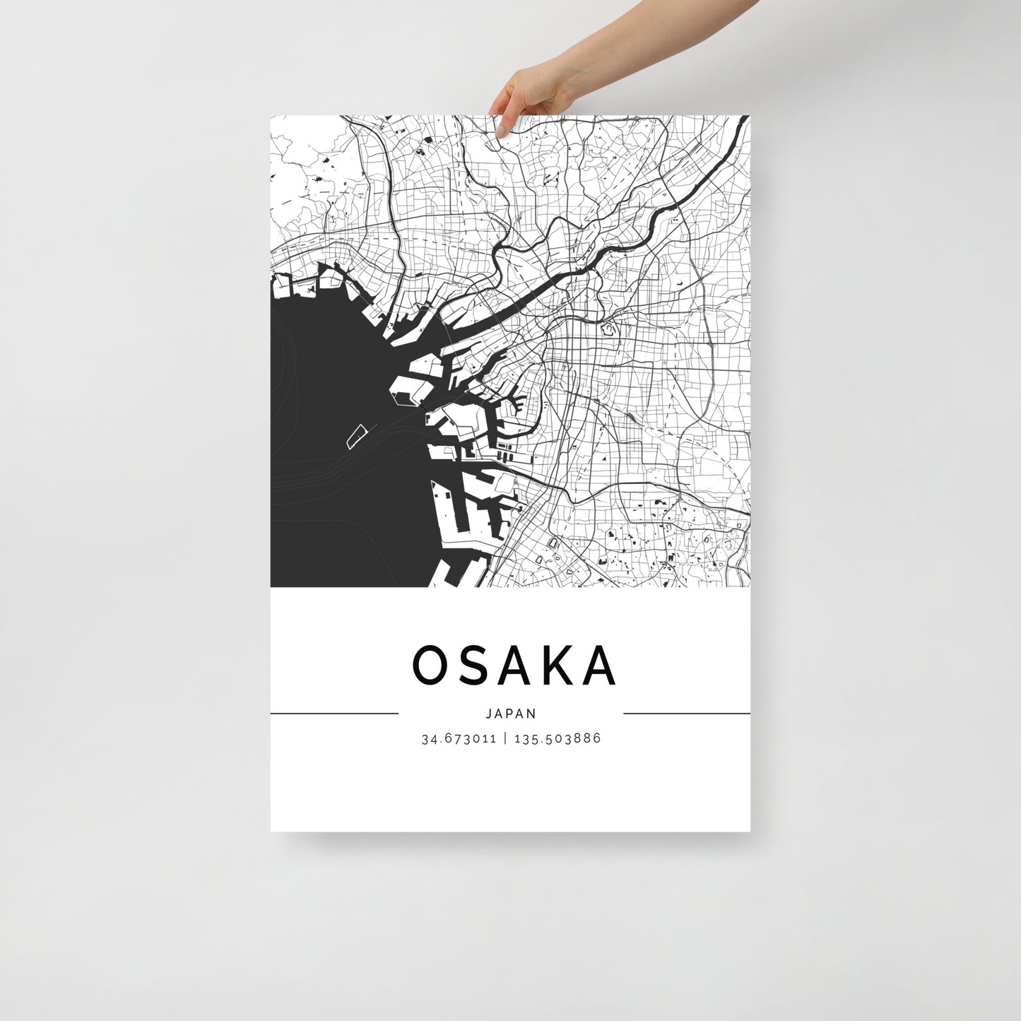 A person holds the Osaka Map by FN Prints, a contemporary black and white poster that elegantly highlights the streets and waterways of Osaka, Japan. This minimalist design serves as stunning wall art, showcasing the coordinates: 34.673011 | 135.503886.