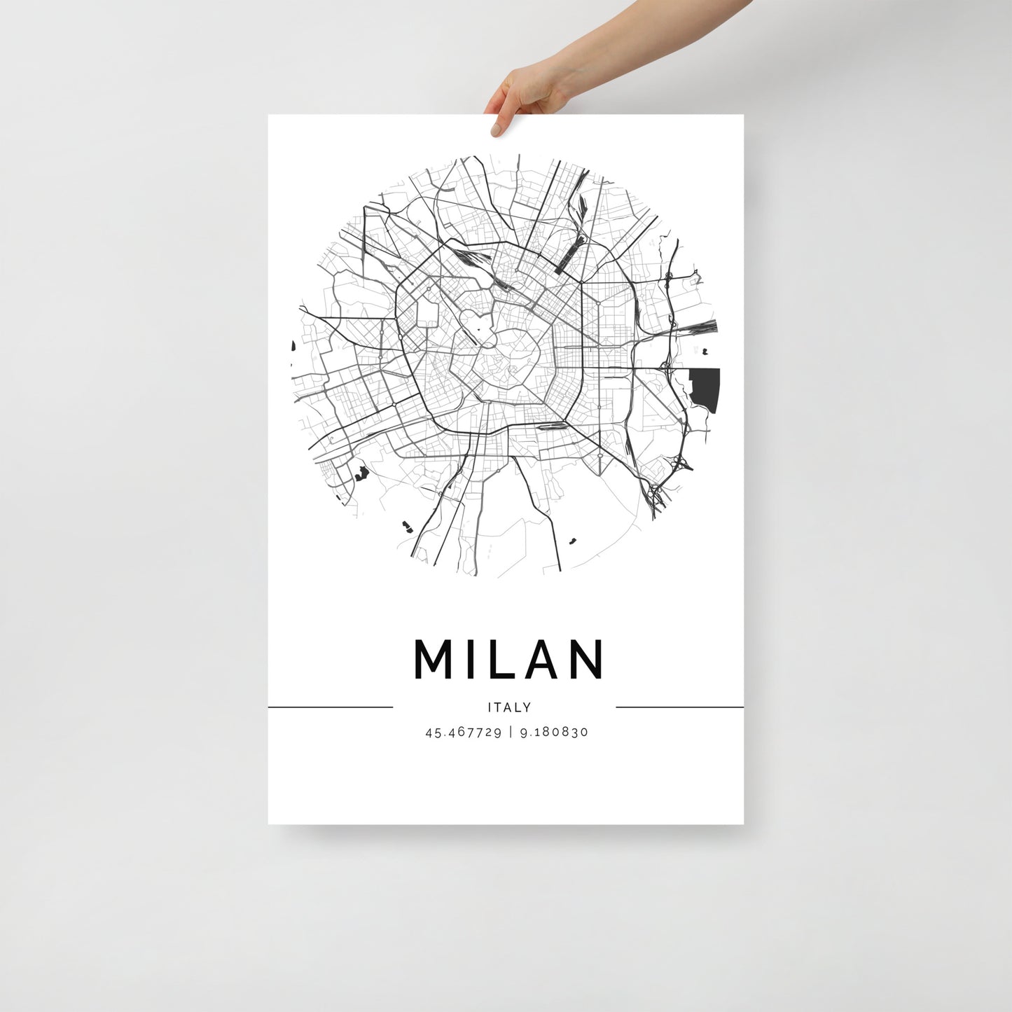 A hand displays a chic wall art piece from FN Prints, showcasing the Milan Map in a sleek black and white circular design. Ideal for urban design enthusiasts, this map features intricate street patterns and coordinates: 45.467729, 9.188030, all with a minimalistic touch.