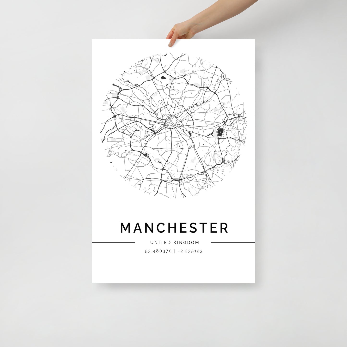 A hand displays the FN Prints Manchester Map poster, featuring a black and white circular street design of the city. Below this piece of urban art, the text reads "MANCHESTER" with its coordinates, 53.480759, -2.242631. The minimalist white background accentuates this stylish wall decor.