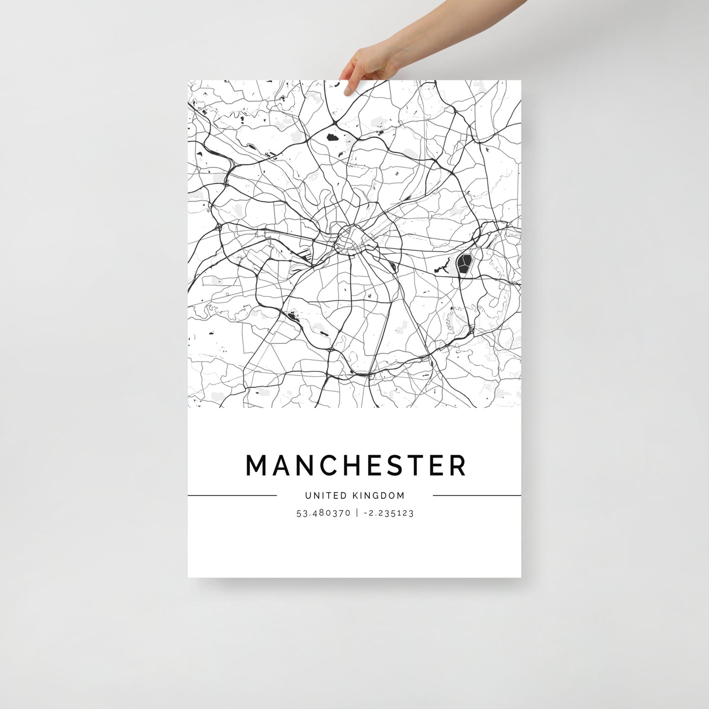 A hand displays a minimalist black and white Manchester Map poster from FN Prints, featuring intricate urban design with detailed road lines and city features. This wall art beautifully presents the coordinates 53.480370, -2.235123 beneath the city's name.