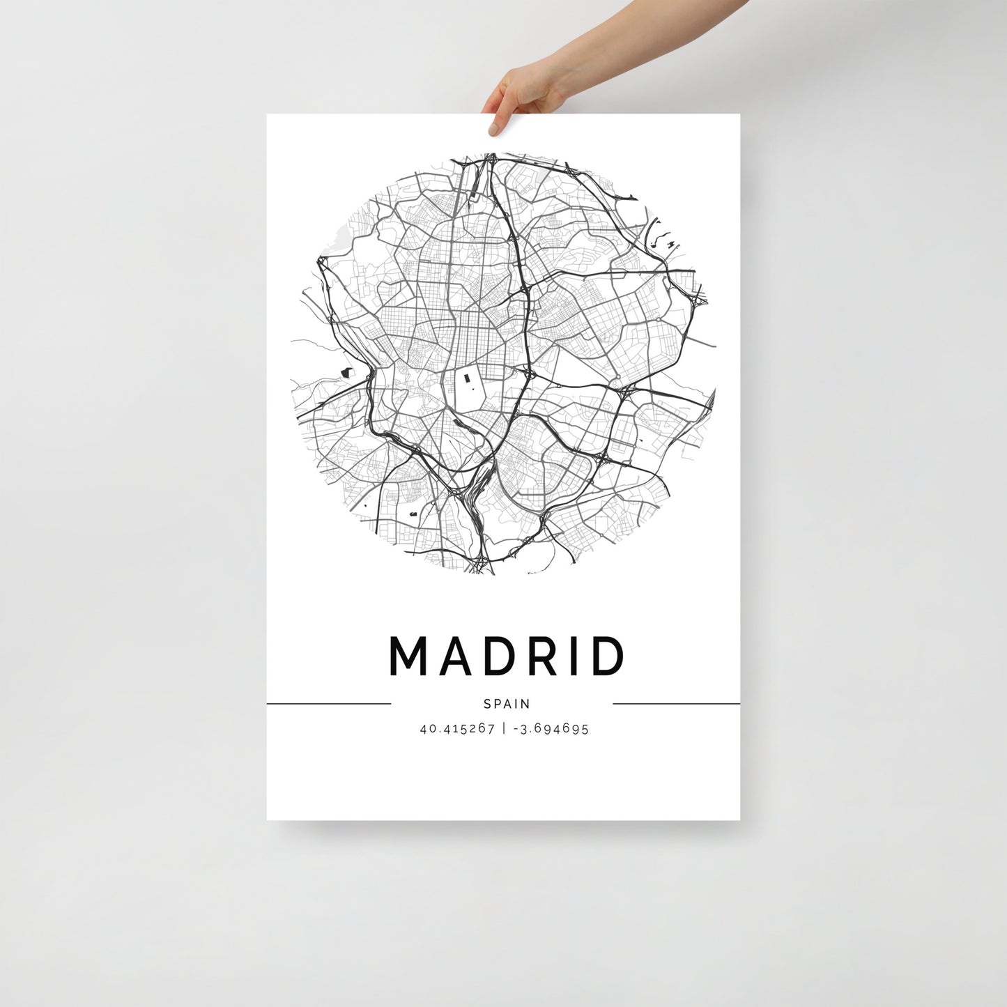 A person is holding a minimalist Madrid Map poster from FN Prints, featuring a circular black and white design. The sleek artwork captures the essence of Spain's capital, elegantly displaying the coordinates 40.4167° N, 3.7046° W at the bottom.