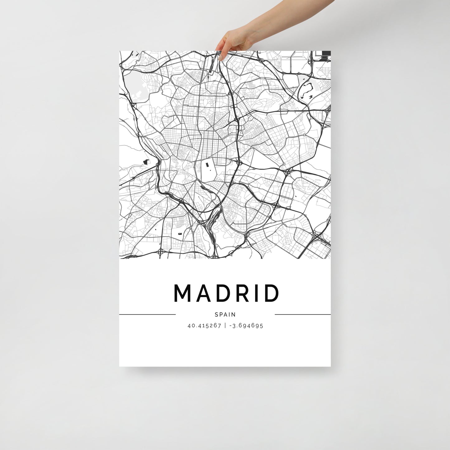 A person holds the FN Prints' Madrid Map, a minimalist black and white poster that features a detailed street map of the city. The artwork elegantly includes the geographic coordinates "40.415287, -3.694665" at the bottom.