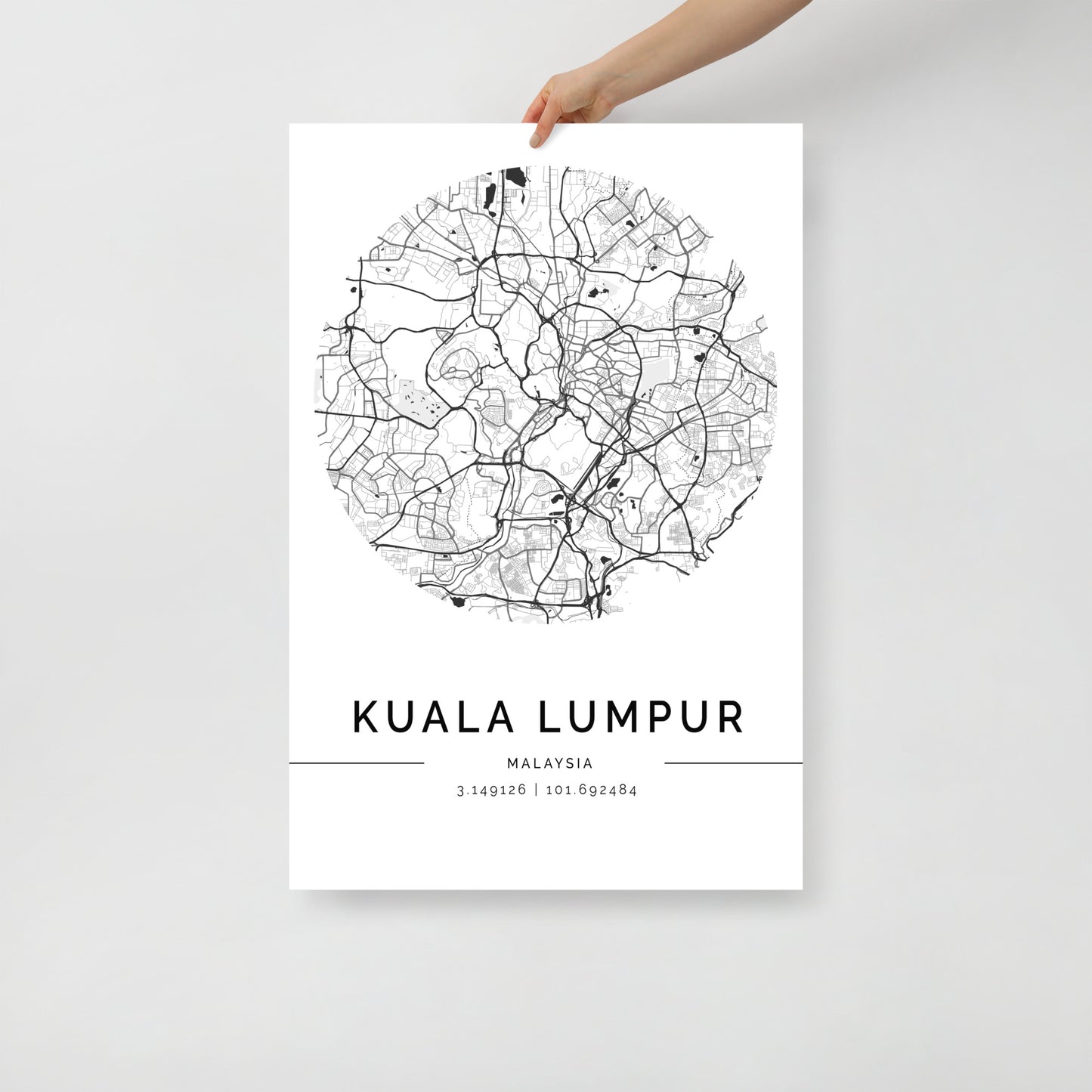 A person is holding the FN Prints' "Kuala Lumpur Map," a minimalist black-and-white poster that intricately captures the city's urban design with detailed street lines in a circular frame. This wall art elegantly showcases Kuala Lumpur's name, country, and precise coordinates: 3.1492°N, 101.6932°E.