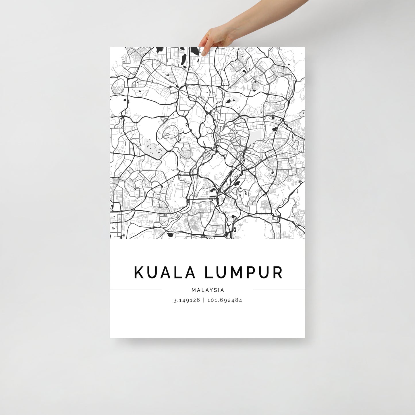 A person is displaying the Kuala Lumpur Map by FN Prints, a minimalist black and white print that highlights its sleek urban design. The geographic coordinates 3.14928° N, 101.69284° E are elegantly printed at the bottom, making it a striking addition to any wall art collection.