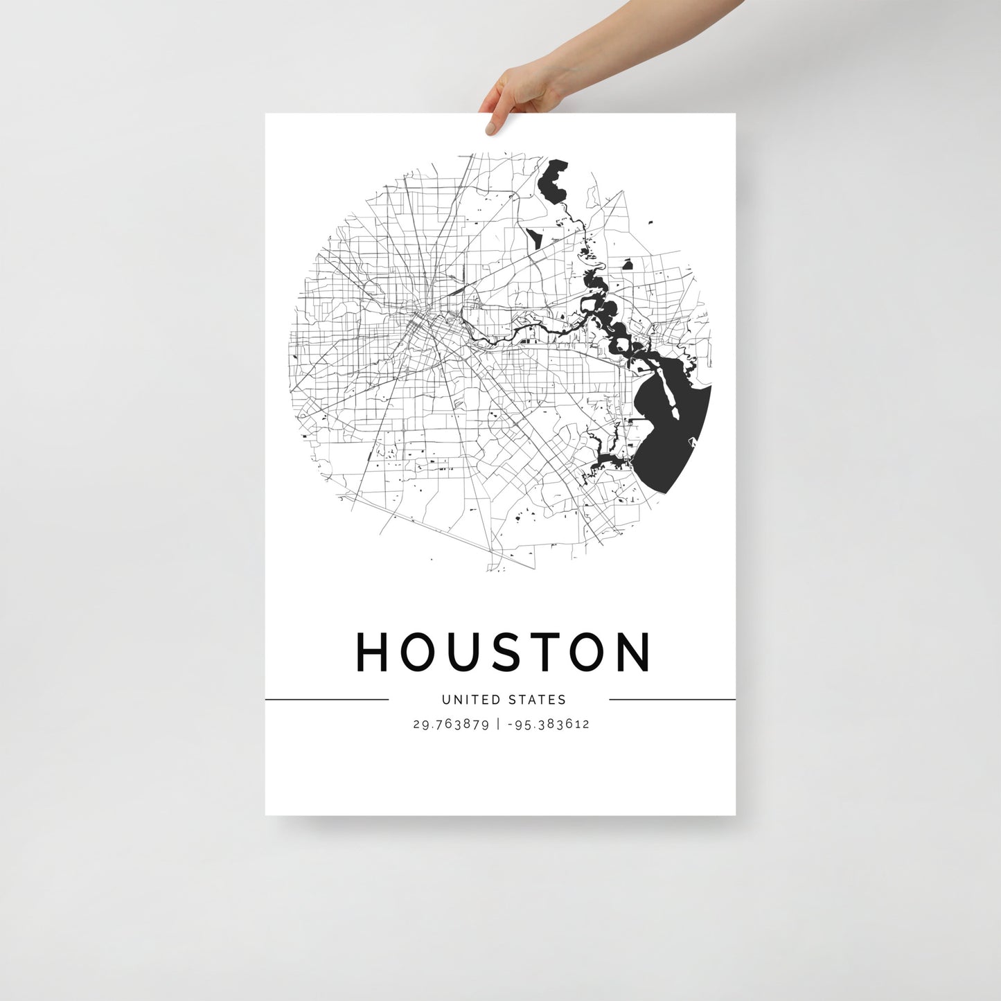 A hand holds the FN Prints Houston Map poster, which showcases a black and white circular design. Below this sophisticated city map wall art are the coordinates 29.763879, -95.383612. The plain white background highlights the artwork's minimalist charm.