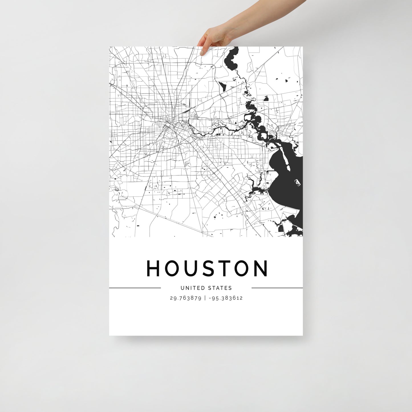 A hand holds an FN Prints Houston Map, featuring the city's intricate streets and waterways in black and white. This eye-catching city map wall art showcases "HOUSTON" along with the exact coordinates, 29.76389° N, 95.38361° W, stylishly presented at the bottom.