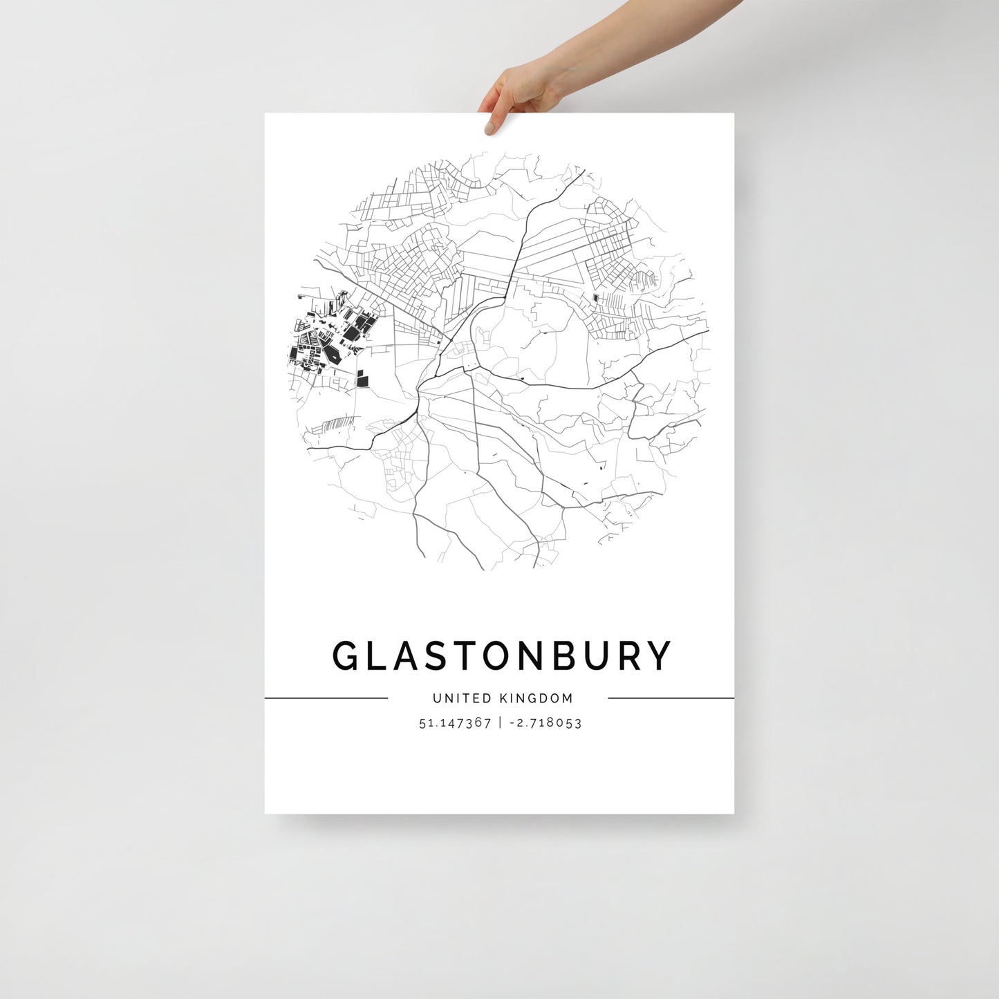 A hand holds up a minimalist Glastonbury Map poster by FN Prints, ideal as wall art. The black and white urban design showcases roads and simple geographical outlines, with the town's name and coordinates displayed below.