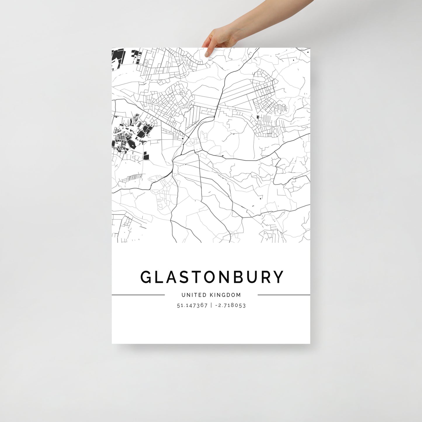 A hand holds an FN Prints Glastonbury Map, a stunning black and white poster that highlights intricate urban design with detailed streets and geography. This elegant piece of wall art includes the coordinates 51.147367, -2.718053 at the bottom, encapsulating the spirit of Glastonbury, United Kingdom.