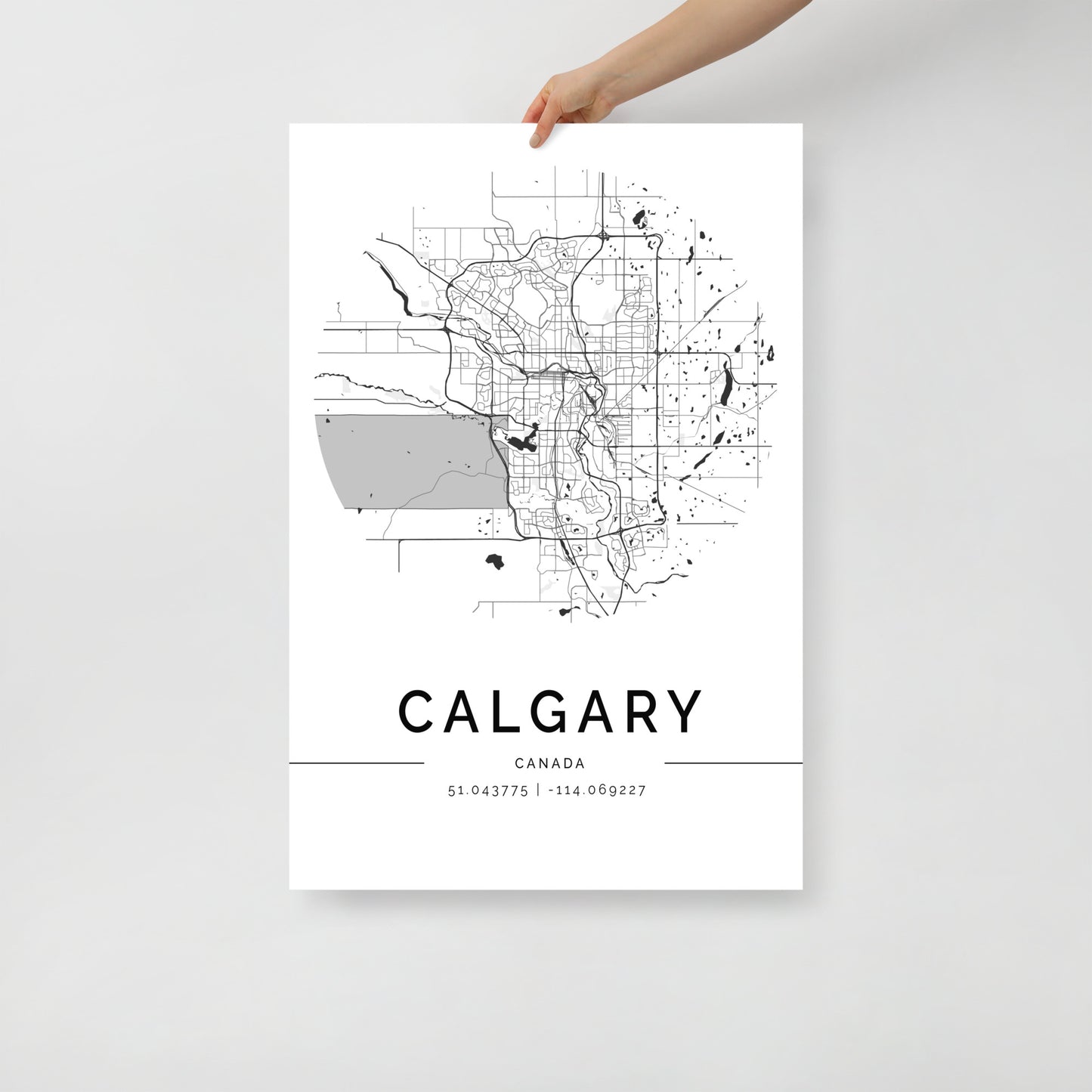 A person holds a black and white FN Prints "Calgary Map" poster, ideal for urban design enthusiasts. This map highlights the city's streets and layout, with the geographic coordinates 51.043775, -114.069227 elegantly displayed below the city name, creating an intriguing piece of wall art.