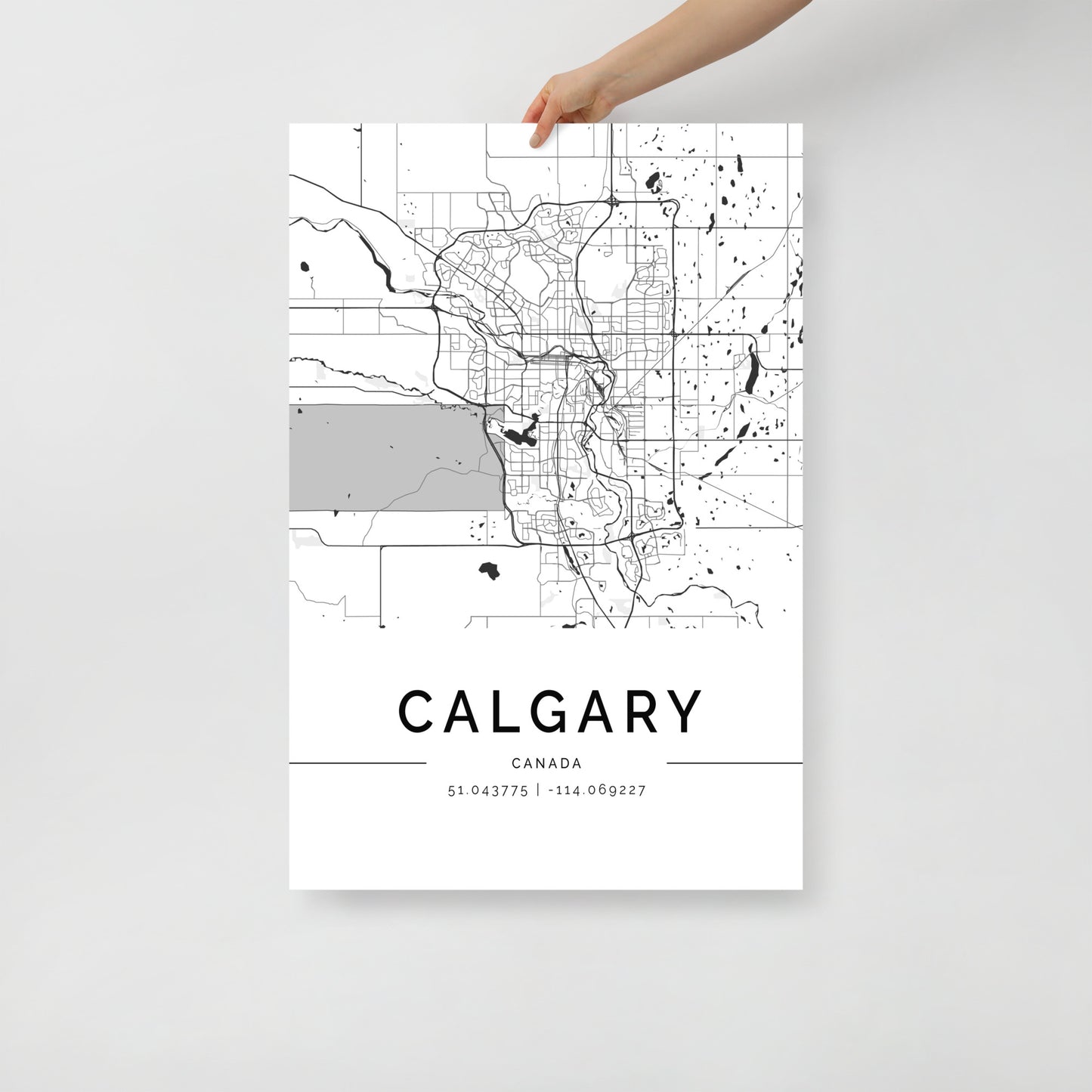 An individual holds a Calgary Map by FN Prints, ideal for urban design enthusiasts. The poster intricately displays streets and city features, along with the coordinates 51.043775, -114.069227 beneath it. This piece of wall art is striking against its simple white background.