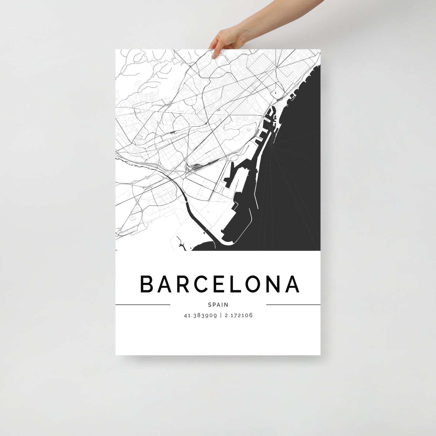 A person's hand holds an FN Prints Barcelona Map, ideal for urban design enthusiasts. This sophisticated wall art showcases a minimalist black and white street grid with the coordinates 41.389909, 2.172106 subtly displayed below.