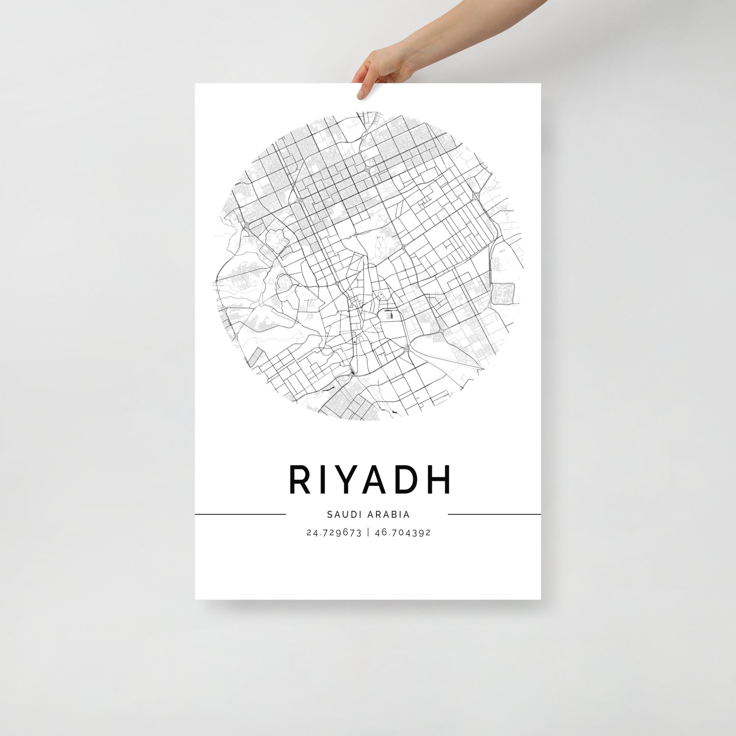 A hand displays an FN Prints Riyadh Map tapestry, showcasing a circular black and white map of Riyadh, Saudi Arabia. Beneath the intricate map are the bold letters "RIYADH" along with the accurate geographical coordinates "24.7190631, 46.704392," all on a simple white background.