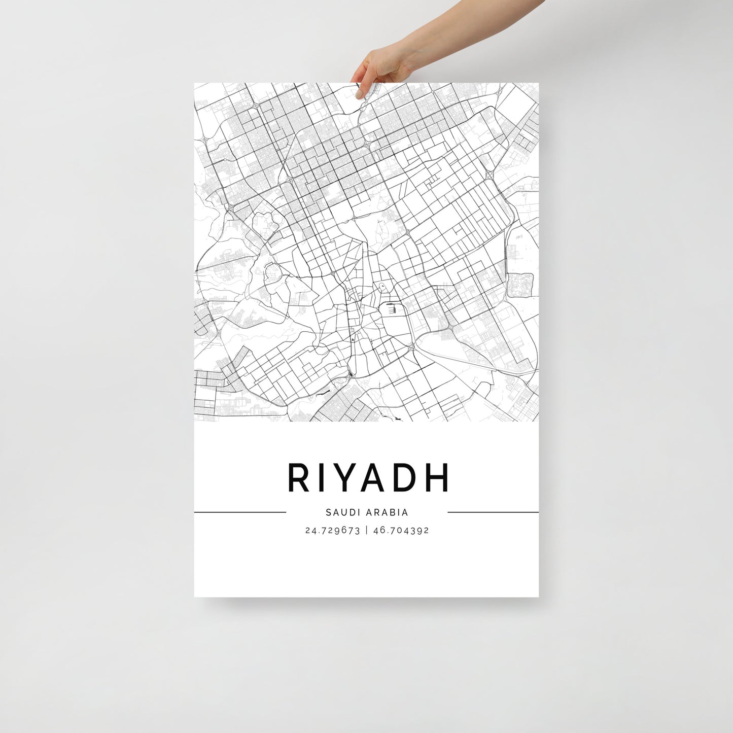 A person's hand holds a FN Prints creation—a Riyadh Map, an intricate black and white urban tapestry. This poster reveals detailed street lines and patterns, elegantly featuring the coordinates 24.729673, 46.704392 at the bottom.