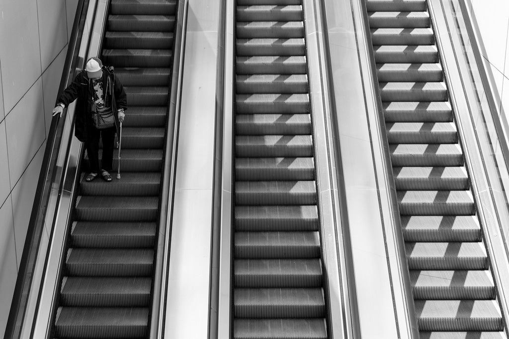 A monochromatic photograph titled "Escalator Ride" by FN Prints captures a person standing and holding onto the railing of an upward-moving escalator. The individual, clad in a hat and jacket while gripping an umbrella, epitomizes urban life art print aesthetics. Parallel escalators on either side create striking black and white contrasts.