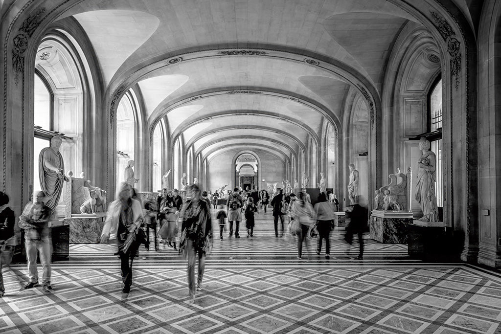 Fine art photography of the interior of the Louvre Museum in Paris, featuring grand vaulted ceilings, stone columns, and classical statues.