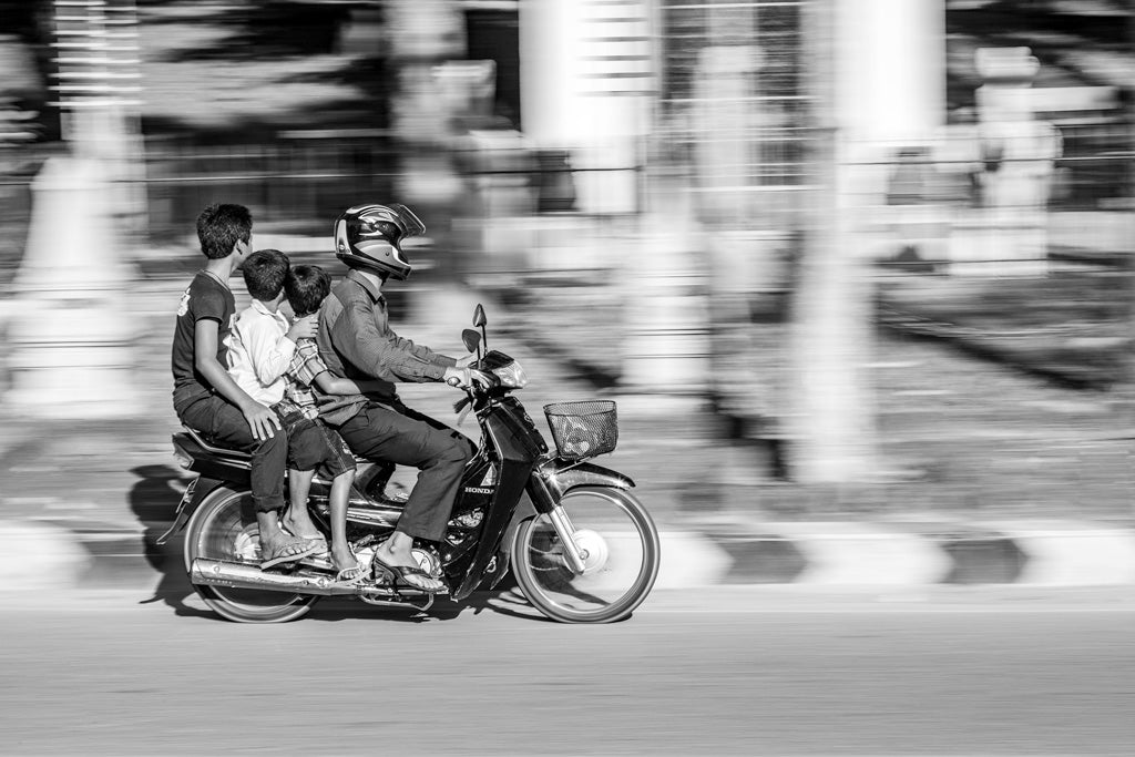 The "Family Journey" photograph from FN Prints captures three children and an adult riding a motorcycle in Phenom Penh. The adult, wearing a helmet, drives as the children sit behind without helmets. The use of long exposure technique creates a blurred background that highlights their speed and adds a dynamic feel to the image.