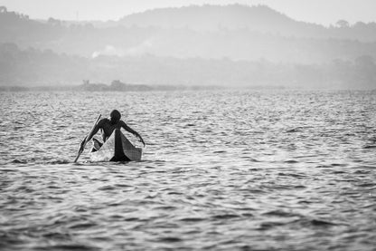 A person is paddling a small boat on a large body of water, holding a net. The scene, reminiscent of Lake Victoria photography, is in black and white with distant mountains and a hazy sky in the background. The calm water enhances the serene atmosphere, making "Fisherman's Journey" by FN Prints perfect for any wall art collection.