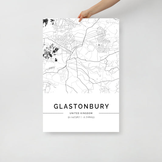 A person holds up the Glastonbury City Map by FN Prints, styled as a black and white map print. This elegant wall art showcases detailed streets and geographic elements, with "GLASTONBURY UNITED KINGDOM" inscribed above the coordinates "51.147367 | -2.718053" beneath the intricate design.