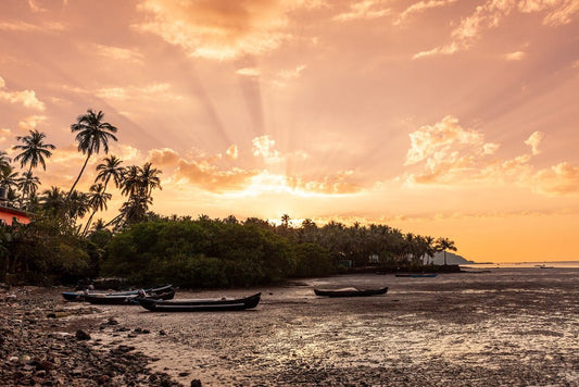 The "Goan Beach Sunset" by FN Prints features a tranquil tropical sunset with golden-orange skies. Sun rays gently filter through the clouds, casting a warm glow on palm trees and a shoreline dotted with moored boats. This photograph beautifully captures the calm reflection of the warm sky on the water, creating a serene and picturesque view brimming with coastal charm.