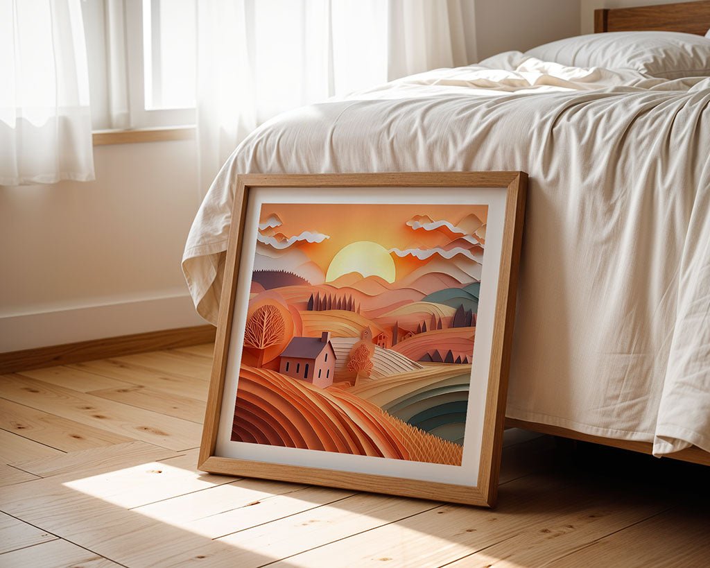 The "Golden Countryside Sunset" digital art piece by FN Prints, featuring a framed depiction of hills, a small house, and trees under a sunset sky, rests on a wooden floor against a bed draped in white sheets. Sunlight streaming through the window casts gentle shadows that highlight the folded paper effect in the artwork.