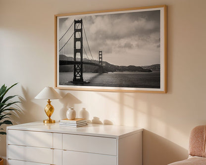 A fine art photograph titled "Golden Gate Bridge" by FN Prints hangs on a beige wall above a white dresser. The dresser is adorned with a small lamp, a vase, and stacked books, while sunlight filters into the room, creating soft shadows.