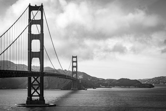 A black and white fine art piece from FN Prints captures the iconic architecture of their "Golden Gate Bridge" photograph, showcasing the bridge gracefully spanning over the water. In the background, an overcast sky with scattered clouds looms, complemented by distant hills and a touch of shoreline to complete this breathtaking scene.