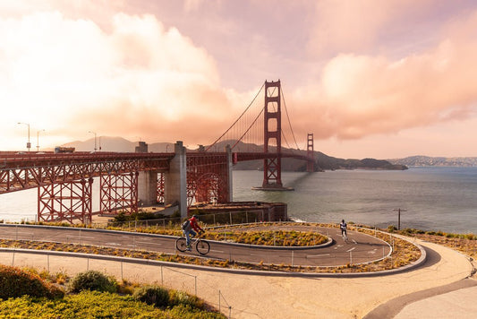 The Golden Gate Cyclist photography print by FN Prints captures the Golden Gate Bridge in San Francisco under a sky with gentle pink and orange clouds. In the foreground, cyclists ride along a winding path, with the iconic red bridge and the bay prominently displayed in the background.