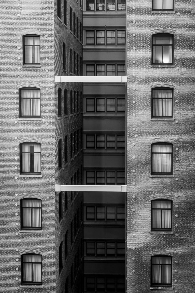 A photograph from FN Prints' "High Rise Symmetry" collection captures the urban architecture of a brick building with rows of rectangular windows. The center section is recessed, featuring three horizontal connecting structures between the larger outer sections. Each window has curtains or blinds, emphasizing the high-rise symmetry.