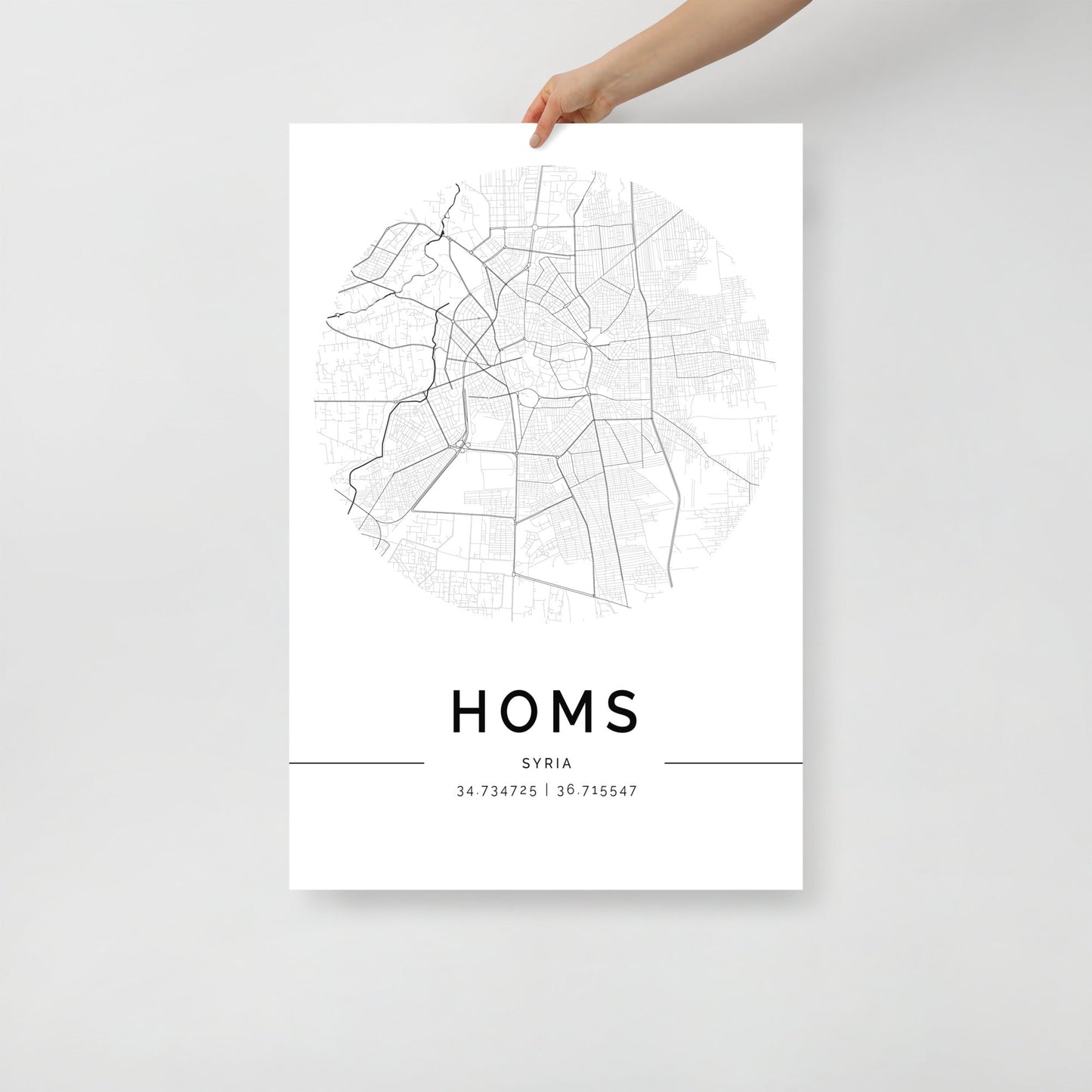 A hand holds a Homs‎ Map by FN Prints, presented in black and white to offer a detailed view of the streets. This minimalist wall art print elegantly features the coordinates "34.734725 | 36.715547" below, making it a timeless addition to any space.