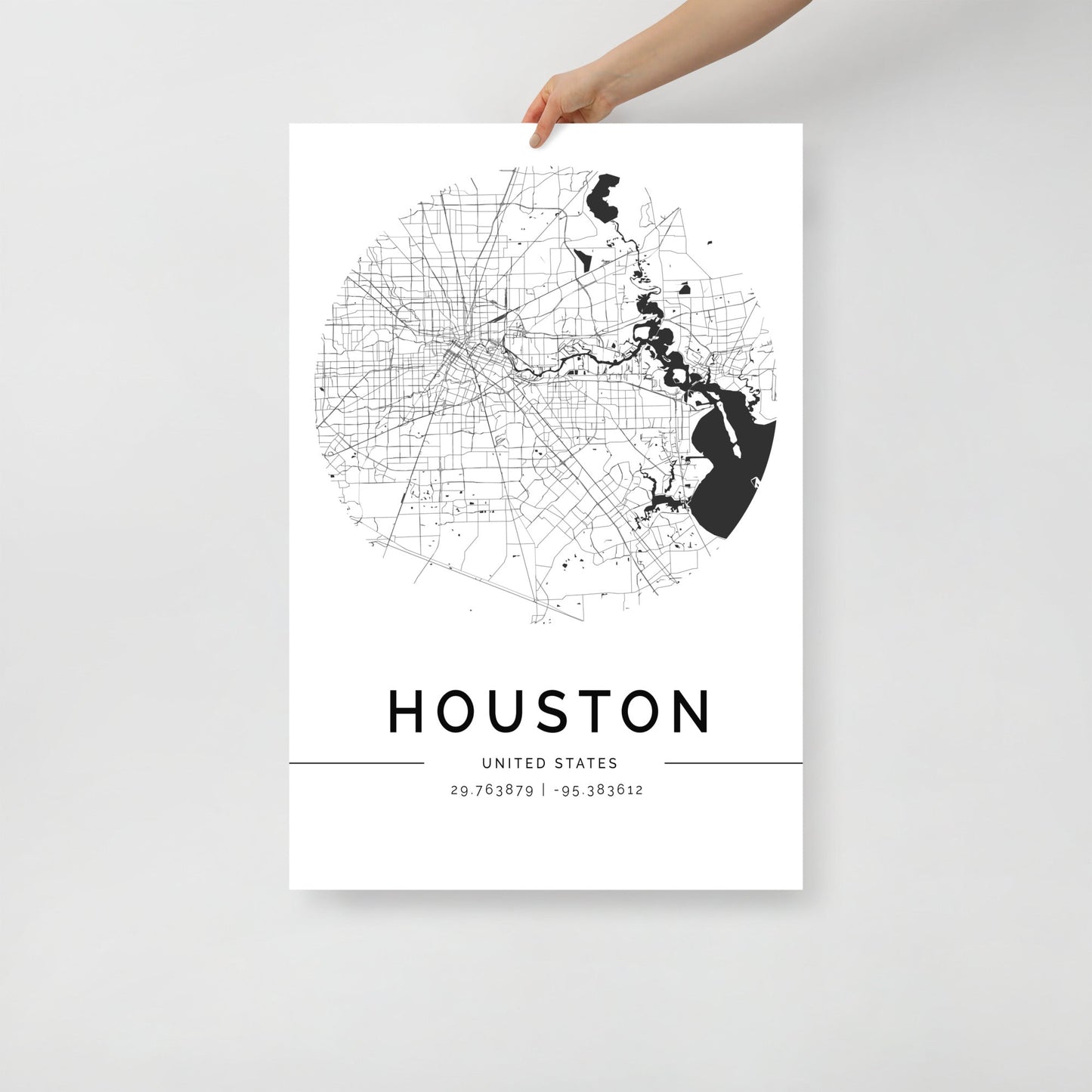 An individual holds the FN Prints "Houston City Map" poster, showcasing a circular, black and white design. It intricately details roads and geographical elements. The word "HOUSTON" along with the coordinates "29.763879, -95.383612" adorns the bottom, making it an ideal piece of wall art for any space.