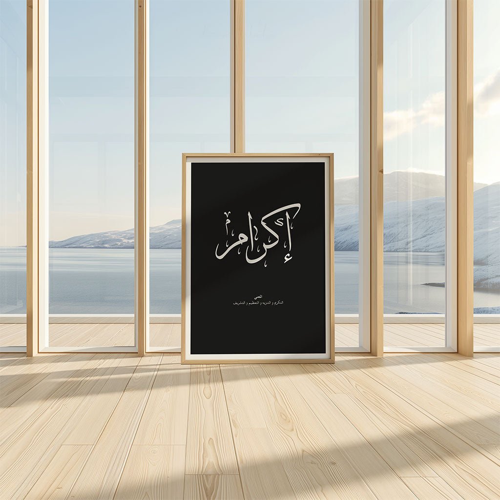 The Ikram - إكرام wall art by FN Prints, featuring Arabic calligraphy on museum-grade paper, stands framed on a light wooden floor near glass windows with scenic views of snowy mountains and a lake in a bright, minimal room.