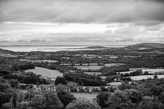 The Irish Landscape Black & White print by FN Prints captures a serene grayscale image of a vast landscape featuring rolling hills, dense forests, and patchwork farmlands that lead to a distant body of water under a cloudy sky. This tranquil scene beautifully showcases Ireland's natural beauty, making it an ideal addition to rustic home decor.