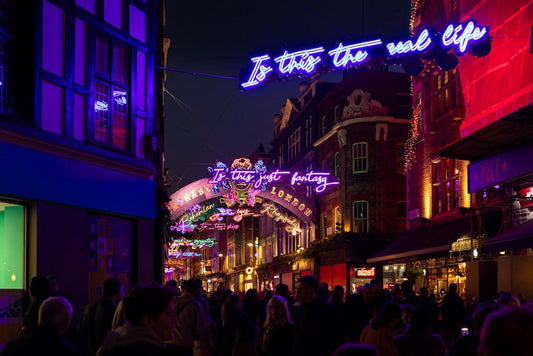 A bustling street scene at night featuring neon signs above, with phrases from FN Prints' "Is This The Real Life" in bright colors. Reminiscent of Carnaby Street, the lively atmosphere is amplified by a festive light display as people walk past vibrant shops.