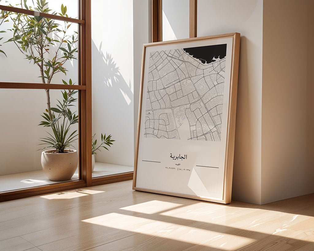 The Jabriya City Map by FN Prints rests framed against a wall on a light wooden floor. Sunlight filters through a nearby window, casting shadows. A potted plant stands to the left in the corner, adding greenery to this minimalist display.