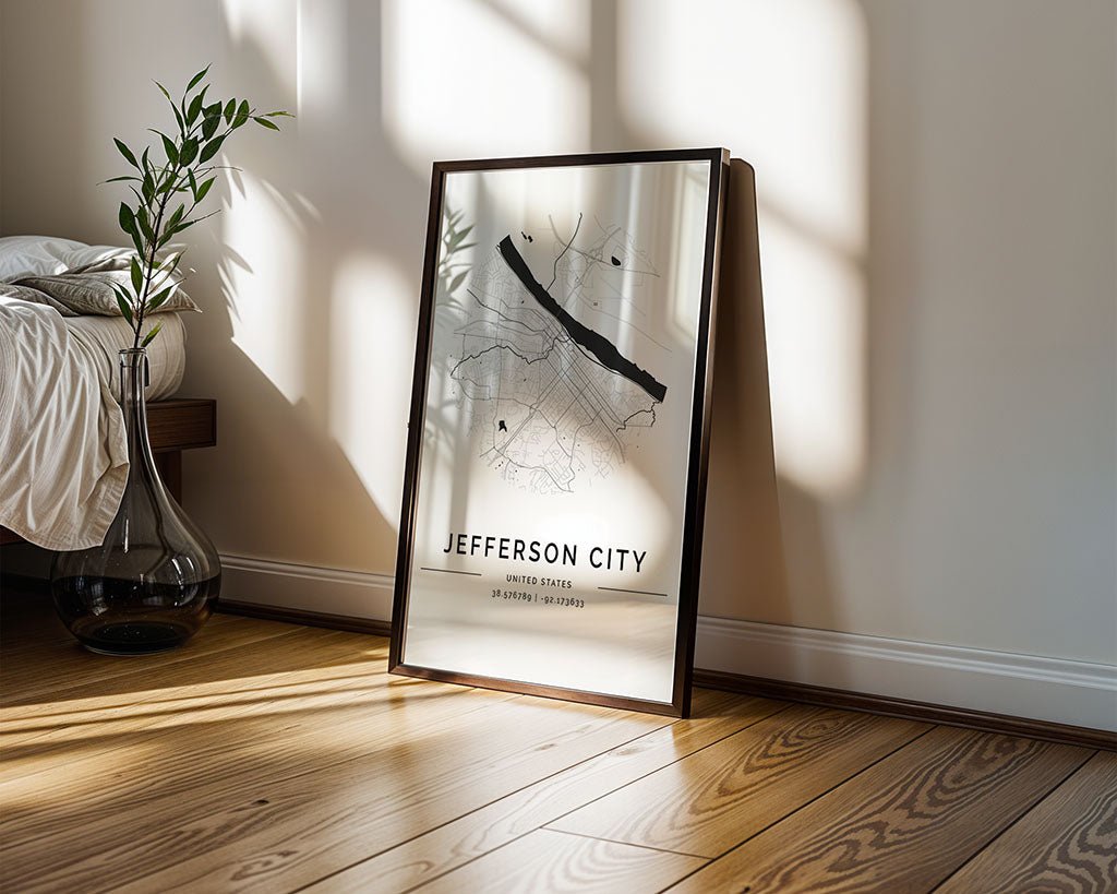 A FN Prints Jefferson City Map, showcasing a minimalist design, leans against the wall on a wooden floor with sunlight casting gentle shadows. A tall clear vase with green branches adds to the serene vibe beside a bed dressed in a white blanket.