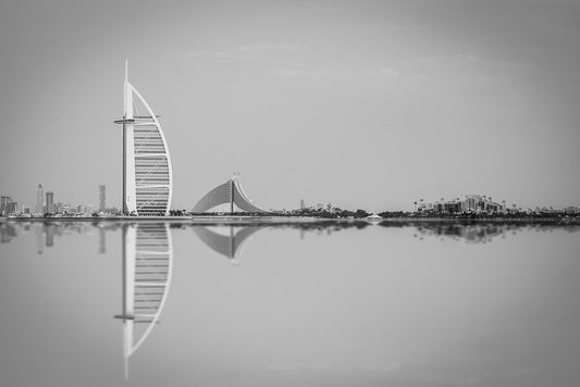 The stunning black and white art photography from FN Prints, titled "Jumeriah Icons," captures the iconic Burj Al Arab hotel in Dubai, UAE. The hotel, with its sail-like structure, sits elegantly on the left beside a smaller building with a curved roof. Both buildings are beautifully reflected in the calm water, highlighting the remarkable architecture of Dubai.