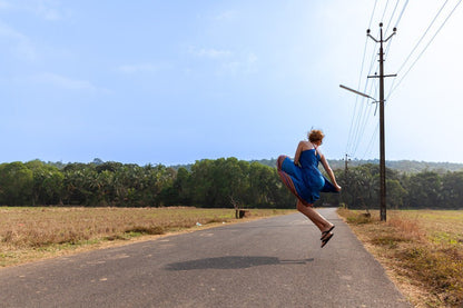 A joyful woman in a blue dress jumps gracefully on an empty rural road, exuding vibrant energy in the "Jumping With Joy" print by FN Prints. The road, framed by open fields and trees beneath a clear blue sky, is complemented by power lines that enhance the picturesque scene, creating a photograph full of natural decor.