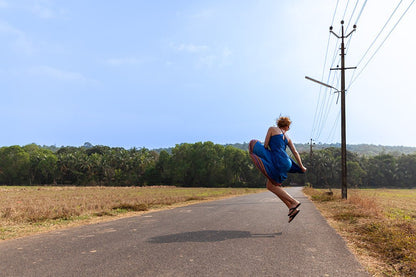 A woman in a blue dress jumping for joy on an empty road lined with tall coconut palm trees in the rural landscape of Goa, India.
