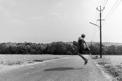 A woman in a blue dress jumping for joy on an empty road lined with tall coconut palm trees in the rural landscape of Goa, India.