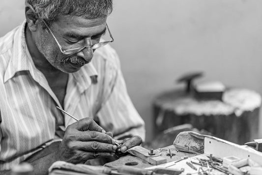 A black-and-white photo captures an elderly man with glasses, carefully handcrafting jewelry at a workbench. He appears focused, holding a small object and a fine tool amidst scattered implements and materials, suggesting the bespoke craftsmanship of Kandy Jeweller by FN Prints in Kandy, Sri Lanka.