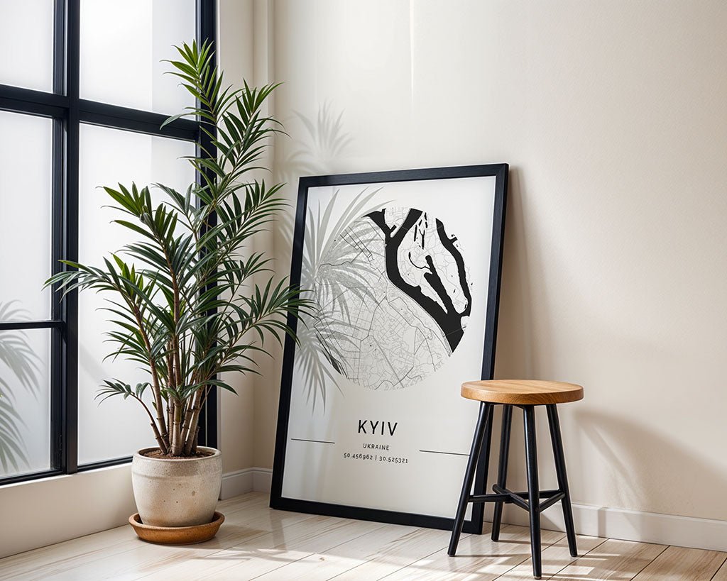 A framed FN Prints Kyiv City Map leans against a light wall beside a tall potted plant, enhancing the minimalist aesthetic. Nearby, a wooden stool with black legs basks in sunlight filtering through a large window, casting gentle shadows on the floor.